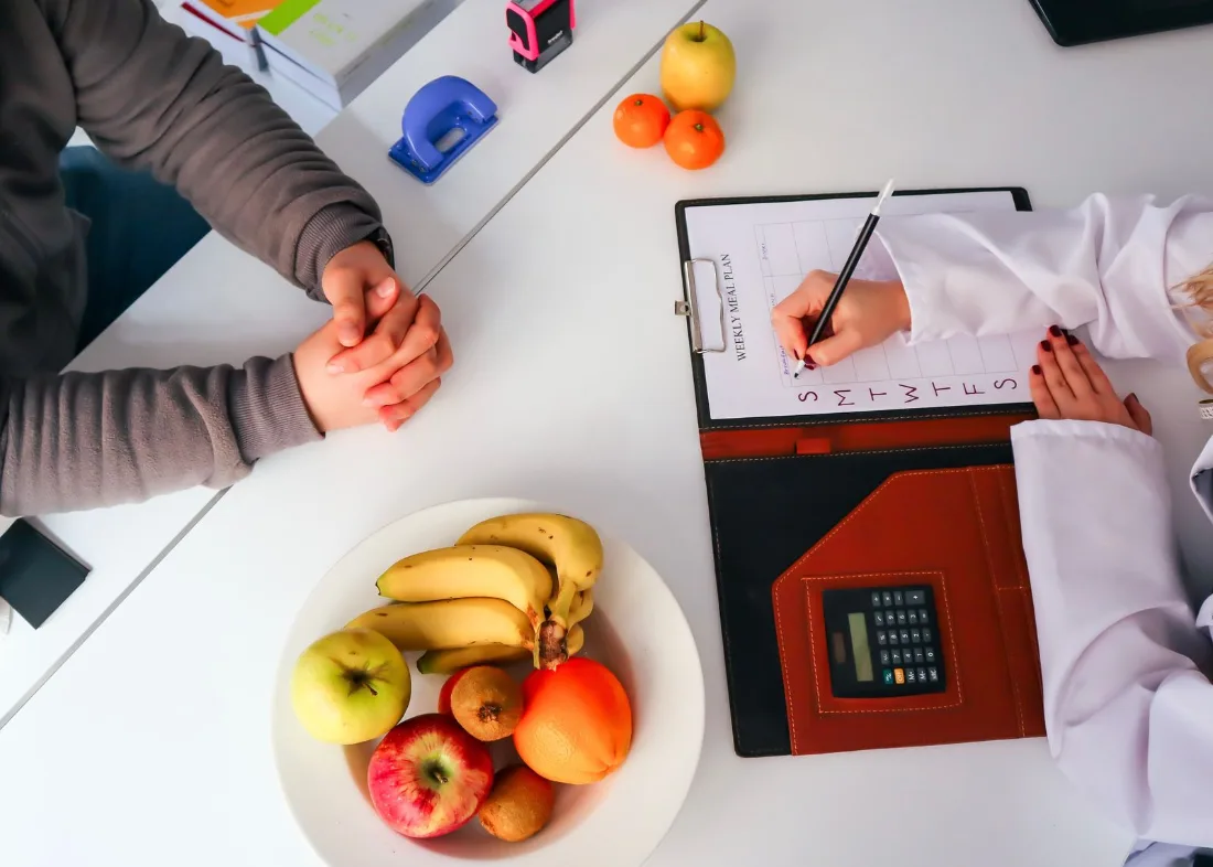 Dietitian writing a weekly meal plan across a desk from a client, with a bowl of fruit between them